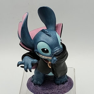 May include: A blue figurine of the character Stitch from Lilo & Stitch, dressed in a black cape and a gold pendant. The figurine has large ears with pink accents and is posed on a purple base.