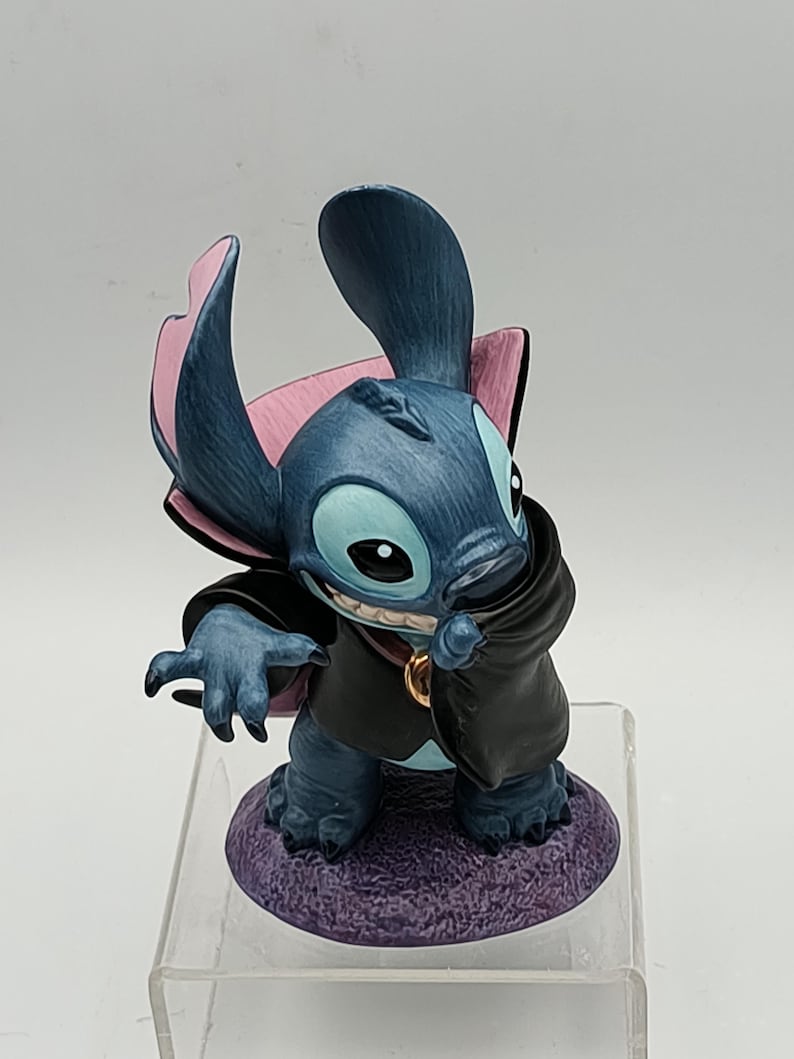 May include: A blue figurine of the character Stitch from Lilo & Stitch, dressed in a black cape and a gold pendant. The figurine has large ears with pink accents and is posed on a purple base.