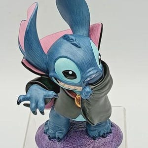 May include: A blue figurine of Stitch, a character from Lilo & Stitch, dressed as a vampire. The figurine has large ears with pink accents, a black cape, and a purple base. The figurine is standing on a clear plastic stand.