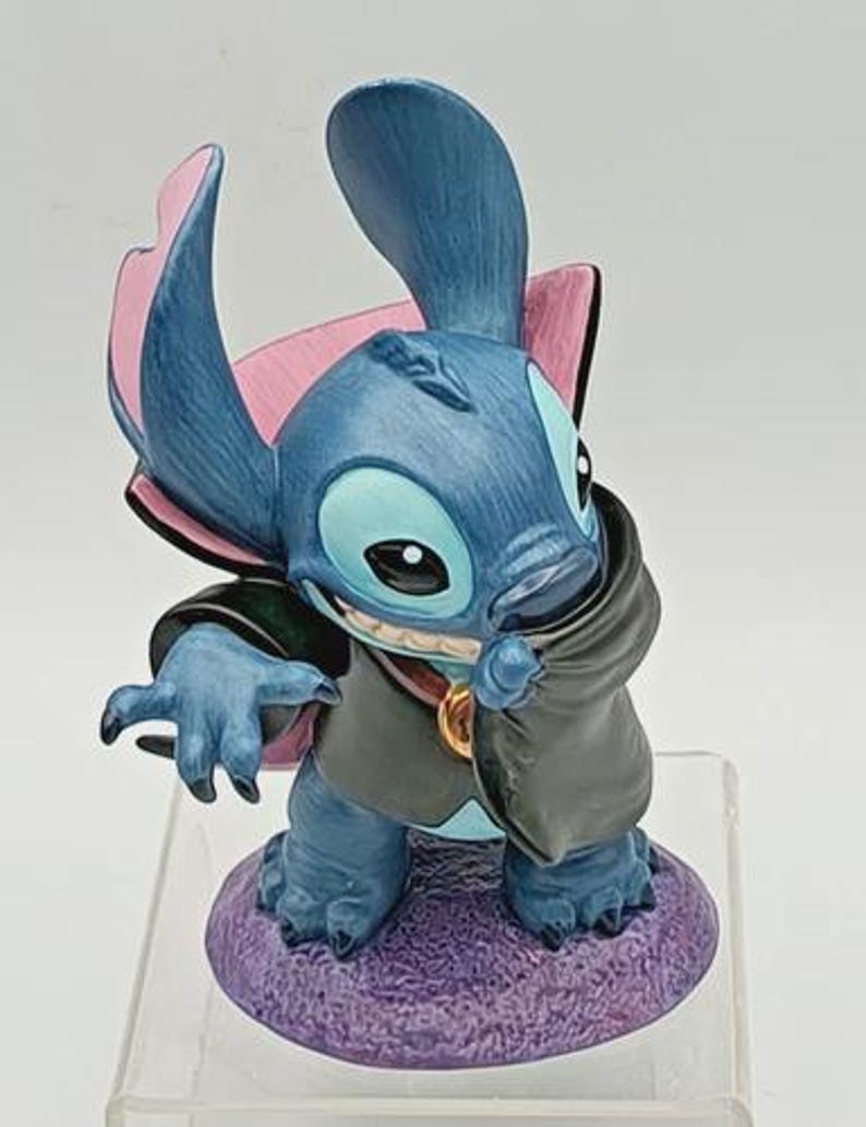 May include: A blue figurine of Stitch, a character from Lilo & Stitch, dressed as a vampire. The figurine has large ears with pink accents, a black cape, and a purple base. The figurine is standing on a clear plastic stand.