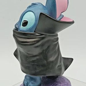 May include: A blue and black figurine of a cartoon character with large ears and a wide open mouth. The figurine is standing on a purple base. The character is wearing a black cape.