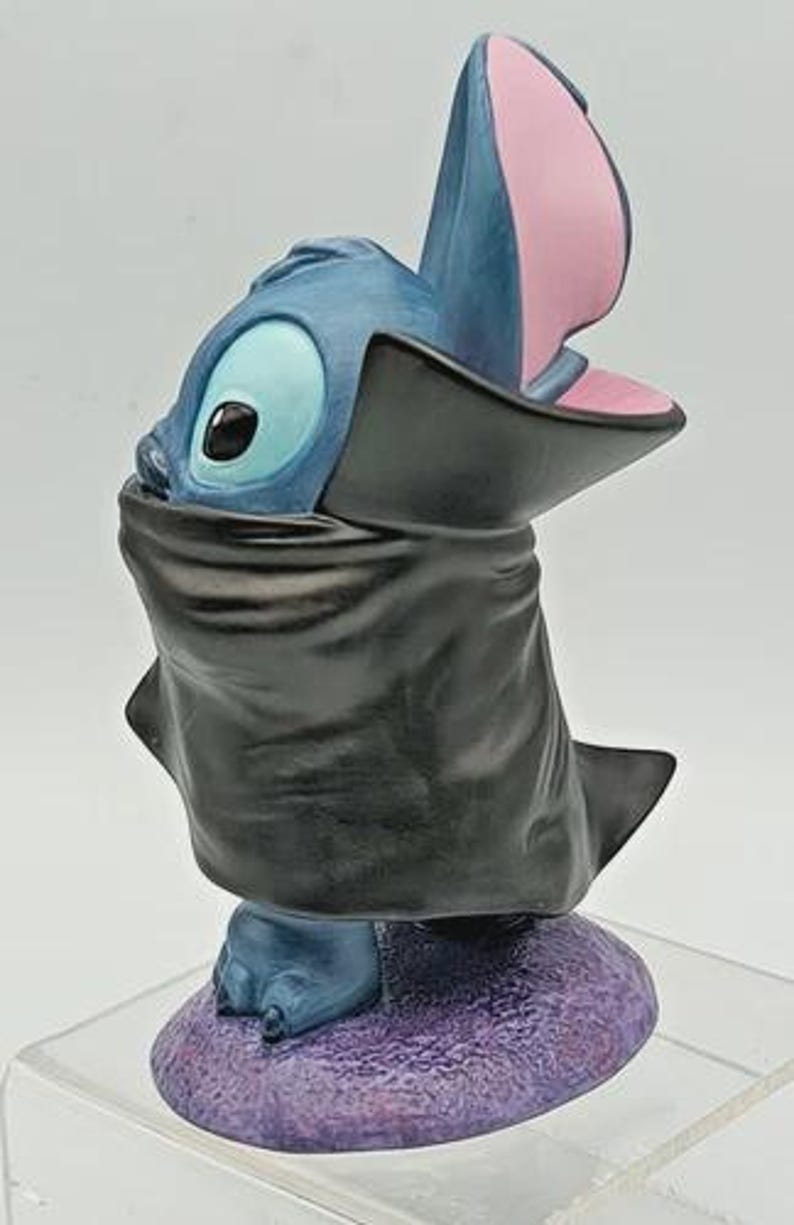 May include: A blue and black figurine of a cartoon character with large ears and a wide open mouth. The figurine is standing on a purple base. The character is wearing a black cape.