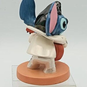 K&ouml;nnte beinhalten: Eine Figur der Disney-Figur Stitch, mit blauem Fell, gro&szlig;en Ohren und einem wei&szlig;en Mantel. Die Figur steht auf einem runden, orangefarbenen Sockel. Die Figur tr&auml;gt einen schwarzen Hut und h&auml;lt ein rotes Objekt.