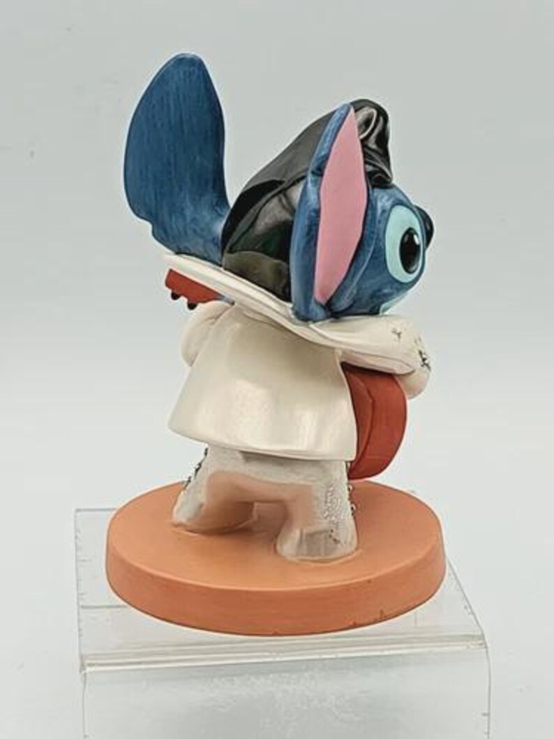 K&ouml;nnte beinhalten: Eine Figur der Disney-Figur Stitch, mit blauem Fell, gro&szlig;en Ohren und einem wei&szlig;en Mantel. Die Figur steht auf einem runden, orangefarbenen Sockel. Die Figur tr&auml;gt einen schwarzen Hut und h&auml;lt ein rotes Objekt.