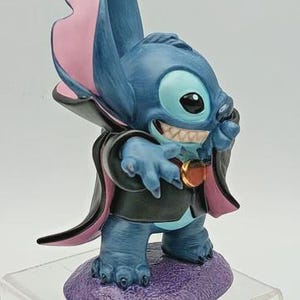 May include: A blue figurine of the character Stitch, dressed in a vampire costume. The figurine has large ears with pink insides, a black cape with pink lining, and a purple base. The figurine is smiling, showing its teeth.
