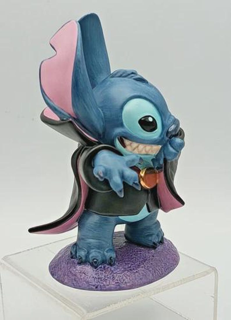 May include: A blue figurine of the character Stitch, dressed in a vampire costume. The figurine has large ears with pink insides, a black cape with pink lining, and a purple base. The figurine is smiling, showing its teeth.