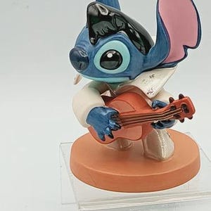 K&ouml;nnte beinhalten: Eine blaue und schwarze Stitch-Figur, die Ukulele spielt. Die Figur hat gro&szlig;e blaue Augen, rosa Ohren und tr&auml;gt ein wei&szlig;es Hemd. Die Figur steht auf einem runden, orangefarbenen Sockel.