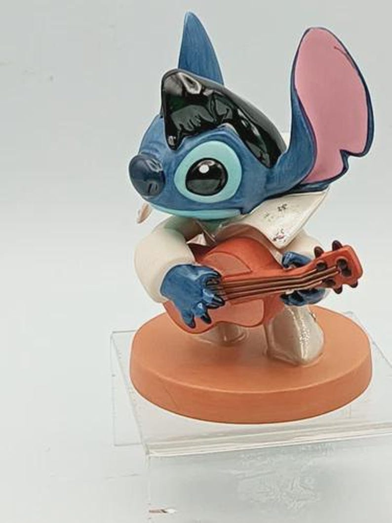 K&ouml;nnte beinhalten: Eine blaue und schwarze Stitch-Figur, die Ukulele spielt. Die Figur hat gro&szlig;e blaue Augen, rosa Ohren und tr&auml;gt ein wei&szlig;es Hemd. Die Figur steht auf einem runden, orangefarbenen Sockel.