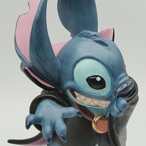 May include: A blue figurine of the character Stitch, with large ears, a mischievous grin, and a black cape. The figurine has pink accents on the ears and a gold-colored medallion.