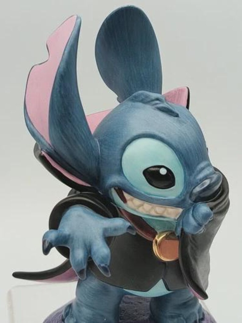 May include: A blue figurine of the character Stitch, with large ears, a mischievous grin, and a black cape. The figurine has pink accents on the ears and a gold-colored medallion.