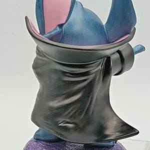 May include: A figurine of a blue alien-like character with large ears and a black cloak. The ears are blue on the outside and pink on the inside. The figurine is standing on a purple base.