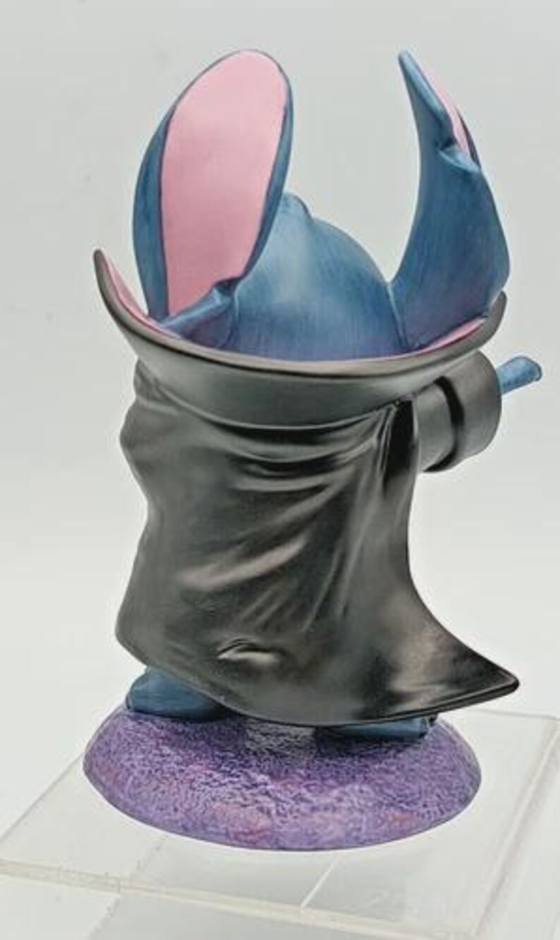 May include: A figurine of a blue alien-like character with large ears and a black cloak. The ears are blue on the outside and pink on the inside. The figurine is standing on a purple base.