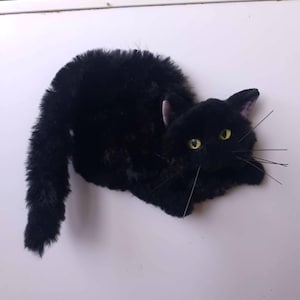 May include: A plush black cat toy with long, shaggy fur, bright yellow eyes, and thin black whiskers. The cat is depicted in a lying position with its tail curved upward. The ears have a subtle purple tint on the inner side, and the overall texture is soft and fuzzy.