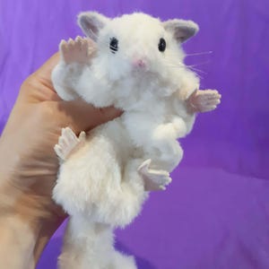 May include: A fluffy white hamster toy with grey ears and pink paws and nose. The toy is held up against a purple background. The hamster has black eyes and long whiskers.