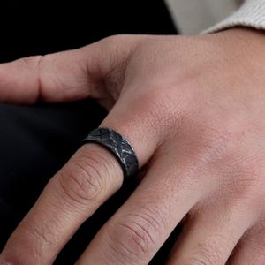 May include: A black ring with a textured, patterned design. The ring is worn on a person's finger.