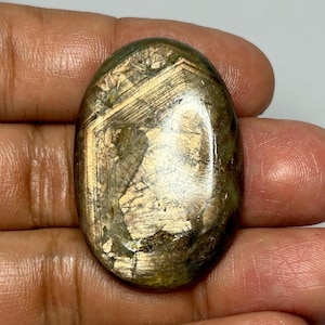 Op de afbeelding: Een ovale edelsteen met goud- en olijfgroene tinten. Het gepolijste oppervlak toont interne patronen en lijnen. De steen wordt in een hand gehouden, wat de grootte en vorm benadrukt. Geschikt voor het maken van sieraden.