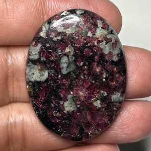 Peut inclure: Un cabochon de pierre précieuse poli de forme ovale présentant un motif tacheté d'inclusions minérales rouge magenta profond, blanc opaque et noir. La surface est lisse et réfléchissante, révélant une texture cristalline naturelle.