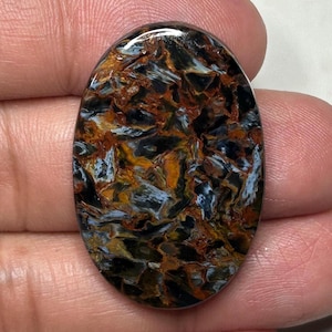 Puede incluir: Gema ovalada con un llamativo patrón de negro, marrón, naranja y blanco. La superficie pulida de la piedra muestra una apariencia compleja y veteada, con los colores arremolinándose. La gema se sostiene en una mano, mostrando su tamaño y detalle.
