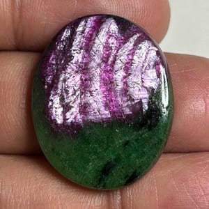 May include: An oval-shaped cabochon gemstone featuring a vibrant magenta-pink crystalline section at the top and a solid, opaque forest green base. The surface is polished and smooth, displaying a distinct color contrast between the two mineral zones.