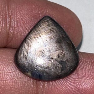 Könnte beinhalten: Ein birnenförmiger, polierter Edelstein mit metallisch silbergrauer Optik. Die Oberfläche weist ein Muster aus sich kreuzenden Linien und subtilen Farbvariationen auf. Der Edelstein wird zwischen zwei Fingern gehalten, wodurch seine Form und Textur hervorgehoben werden.