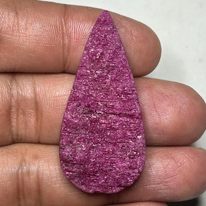 May include: A teardrop-shaped gemstone with a vibrant magenta hue and a textured, crystalline surface. The stone features a pointed top and a rounded base, displaying a rough, natural appearance with visible sparkling facets across its face.