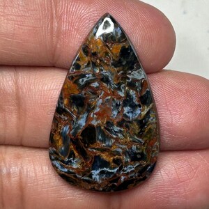 May include: A teardrop-shaped gemstone with a polished surface. The stone features a complex pattern of swirling colors, including shades of brown, black, blue, and gold. The gemstone is held in a hand, showcasing its size and shape.