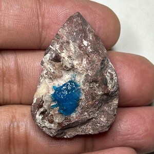 Puede incluir: Una roca rugosa en forma de lágrima con un depósito mineral azul vibrante. La superficie de la roca es una mezcla de tonos marrones, beige y blancos, creando una apariencia texturizada. El mineral azul brillante contrasta fuertemente con los tonos terrosos de la piedra.
