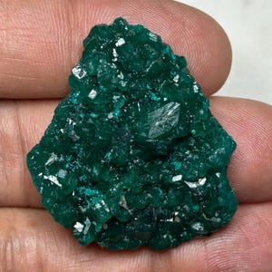 Könnte beinhalten: Ein rohes, tief smaragdgrünes Mineralexemplar, das aus einer Ansammlung kleiner, facettierter Kristalle mit einer reflektierenden, funkelnden Oberfläche besteht. Der Stein hat eine unregelmäßige, gezackte Form mit verschiedenen Grüntönen.