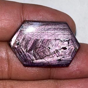 Puede incluir: Una gema hexagonal pulida con un tono púrpura intenso. La piedra presenta un patrón geométrico complejo de líneas y facetas, creando un efecto brillante e iridiscente. La gema se sostiene en una mano.