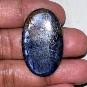 Puede incluir: Gema ovalada con una mezcla de tonos azules, plateados y marrones. La superficie tiene un aspecto texturizado y agrietado, con áreas de azul profundo y plata brillante. La gema se sostiene en una mano.