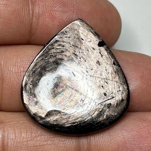 May include: A teardrop-shaped, polished gemstone with a dark, iridescent surface. The stone has a swirling pattern with hints of rainbow colours and fine lines. The gem is held in a hand, showcasing its size and texture.