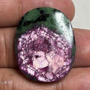 May include: An oval-shaped polished gemstone featuring a vibrant magenta and deep pink center with a crystalline texture, surrounded by a smooth, dark forest green mineral border with black inclusions throughout the stone's surface.