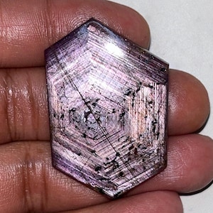 Peut inclure: Une pierre précieuse hexagonale facettée, de couleur rose-violet. La pierre présente une structure interne complexe avec des lignes visibles et des inclusions sombres. La gemme est tenue dans une main, mettant en valeur sa surface polie et son motif unique.