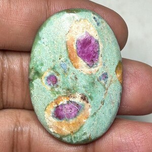 Puede incluir: Una piedra preciosa cabujón de forma ovalada con una superficie lisa y pulida de color base verde claro. La piedra muestra tres inclusiones distintas de color rosa magenta vibrante, cada una rodeada por un suave patrón de halo circular color canela.