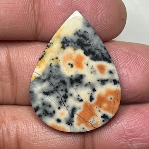 May include: A teardrop-shaped gemstone with a distinctive pattern. The stone displays a blend of cream, black, grey, and orange hues. The surface has a marbled appearance with visible cracks, enhancing its natural appeal. Suitable for crafting jewellery.