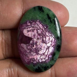Peut inclure: Une pierre précieuse cabochon de forme ovale présentant un centre rose magenta vibrant à la texture cristalline, entouré d'une matrice vert forêt profond avec des inclusions noires éparses, polie pour une finition lisse et brillante.