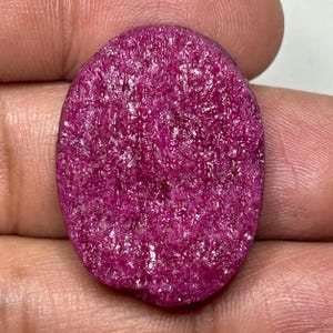 May include: Oval-shaped, rough-cut ruby gemstone. The ruby has a deep, saturated magenta color with a textured surface. The gemstone is held in a hand, showcasing its natural, unpolished appearance. Ideal for jewelry making or gemstone collectors.