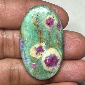 Puede incluir: Una piedra preciosa en forma de cabujón ovalado con una superficie pulida, que presenta un color base verde claro moteado, intercalado con distintas inclusiones de color rosa magenta intenso rodeadas de halos blanco crema y vetas minerales.