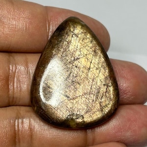 Puede incluir: Una gema pulida en forma de lágrima. La piedra es de color marrón dorado con finas líneas entrecruzadas en su superficie. La gema se sostiene en una mano, resaltando su forma. El objeto parece natural y posiblemente hecho a mano.