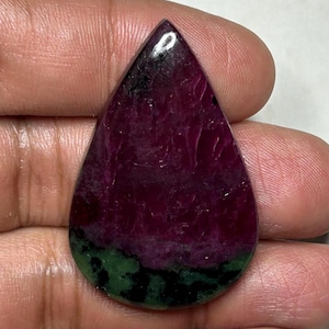Puede incluir: Una piedra preciosa en forma de pera con un corte cabujón que presenta una sección superior en color magenta y púrpura intenso con una base distintiva en verde oscuro y negro. La piedra tiene una superficie pulida con inclusiones.