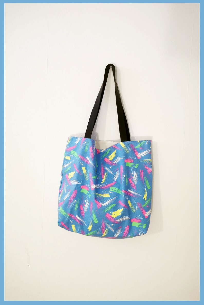 May include: A tote bag featuring a blue base color with a repeating pattern of brushstroke marks in neon pink, bright yellow, lime green, and white. The bag includes two black fabric shoulder straps attached to the top edge of the rectangular body.