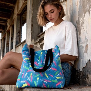 May include: A tote bag featuring a vibrant blue base with a multi-colored abstract brushstroke pattern in shades of pink, yellow, white, and green. The bag includes two sturdy black fabric handles and a visible brown leather-like accent tab.