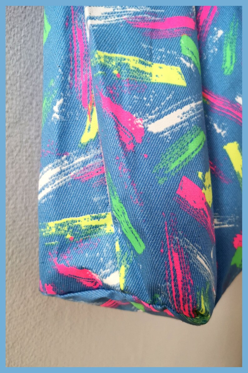 May include: A blue fabric bag featuring a textured, woven surface pattern with diagonal brushstroke designs in vibrant neon pink, bright lime green, and stark white colors, showcasing a repeating abstract paint splatter aesthetic throughout.