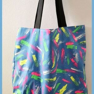 May include: A blue tote bag featuring a repeating pattern of short, thick brushstrokes in neon pink, lime green, and white. The bag has two long, solid black fabric straps attached to the top edge, allowing it to be carried over the shoulder.