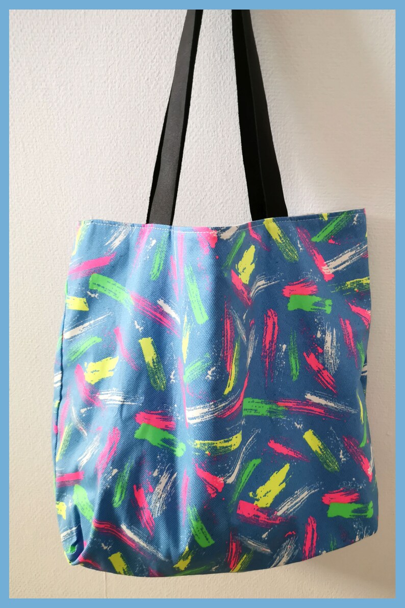 May include: A blue tote bag featuring a repeating pattern of short, thick brushstrokes in neon pink, lime green, and white. The bag has two long, solid black fabric straps attached to the top edge, allowing it to be carried over the shoulder.