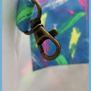 May include: A bronze-toned metal lobster clasp attached to a fabric loop featuring a blue, green, and pink brushstroke pattern. The clasp is connected to a white fabric section on the left and a blue fabric section with colorful paint strokes.