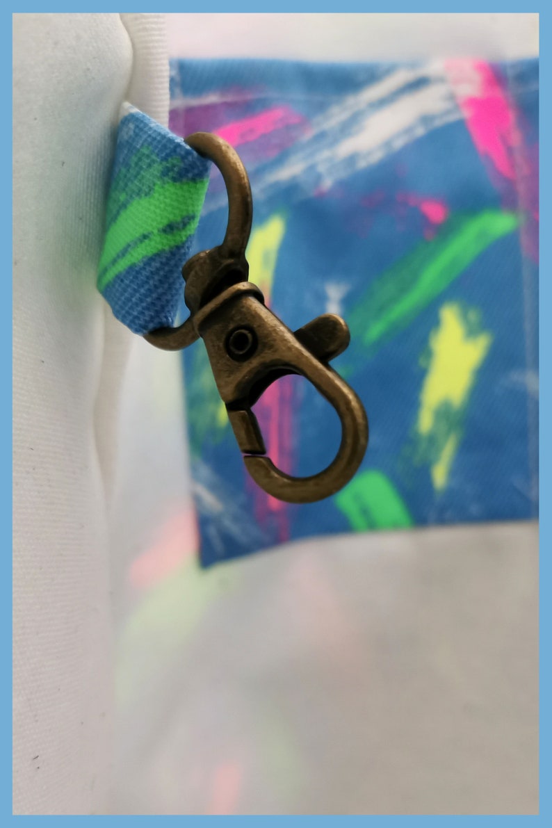 May include: A bronze-toned metal lobster clasp attached to a fabric loop featuring a blue, green, and pink brushstroke pattern. The clasp is connected to a white fabric section on the left and a blue fabric section with colorful paint strokes.