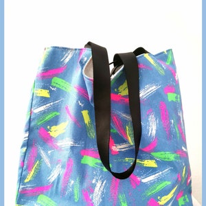 May include: A blue tote bag featuring a repeating pattern of brushstroke marks in neon pink, bright yellow, lime green, and white. The bag has two solid black fabric handles attached to the top, providing a contrast to the colorful patterned body.