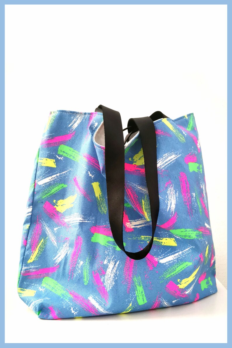 May include: A blue tote bag featuring a repeating pattern of brushstroke marks in neon pink, bright yellow, lime green, and white. The bag has two solid black fabric handles attached to the top, providing a contrast to the colorful patterned body.