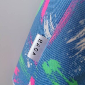 May include: A blue fabric surface featuring a textured pattern of diagonal brushstrokes in bright neon pink, lime green, and white. A small, rectangular white fabric tag is sewn onto the material, displaying the black text BACA. in a sans-serif font.
